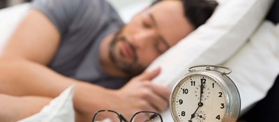 Person asleep with an alarm clock, spectacles and a notepad & pen on a bedside table. Writing down thoughts before bed can help you sleep better