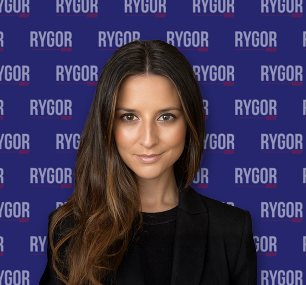 Woman with long brown hair in a black jacket, in front of a blue backdrop repeating the Rygor Group logo.