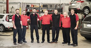 Seven people working in a garage standing in a line with their arms around each other smiling