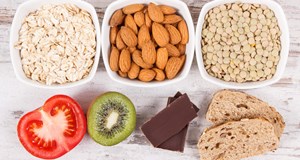 Lots of healthy food is laid out on a wooden table, including bowls of different nuts, fruit, vegetables, dark chocolate and brown bread.