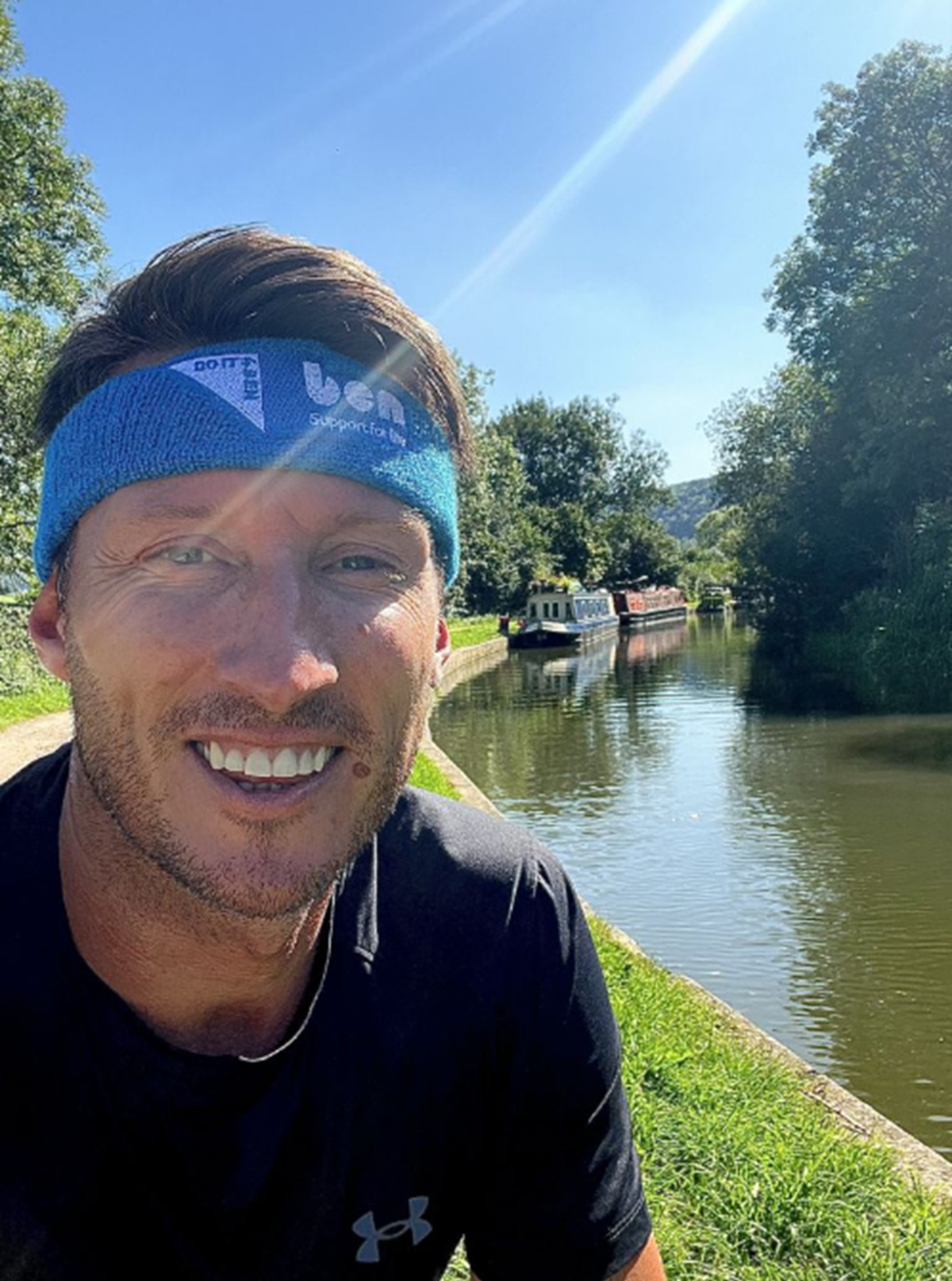 Smiling man wearing a blue Ben 'Support for life' headband taking a selfie on a sunny canal towpath, with a narrowboat moored behind him.