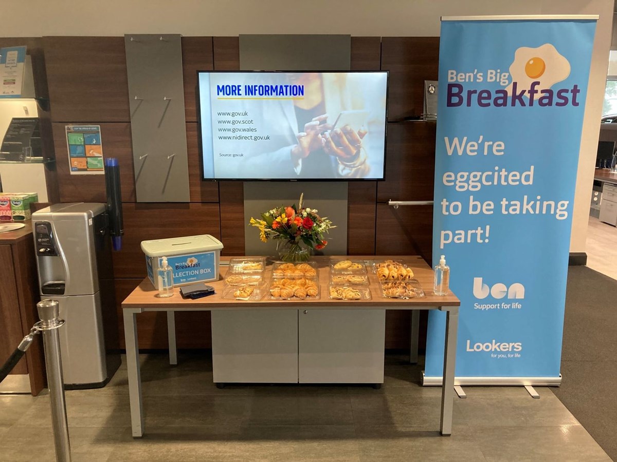 A Ben's Big Breakfast table of pastries and a collection box in a Lookers showroom, beside a banner reading "We're eggcited to be taking part!"