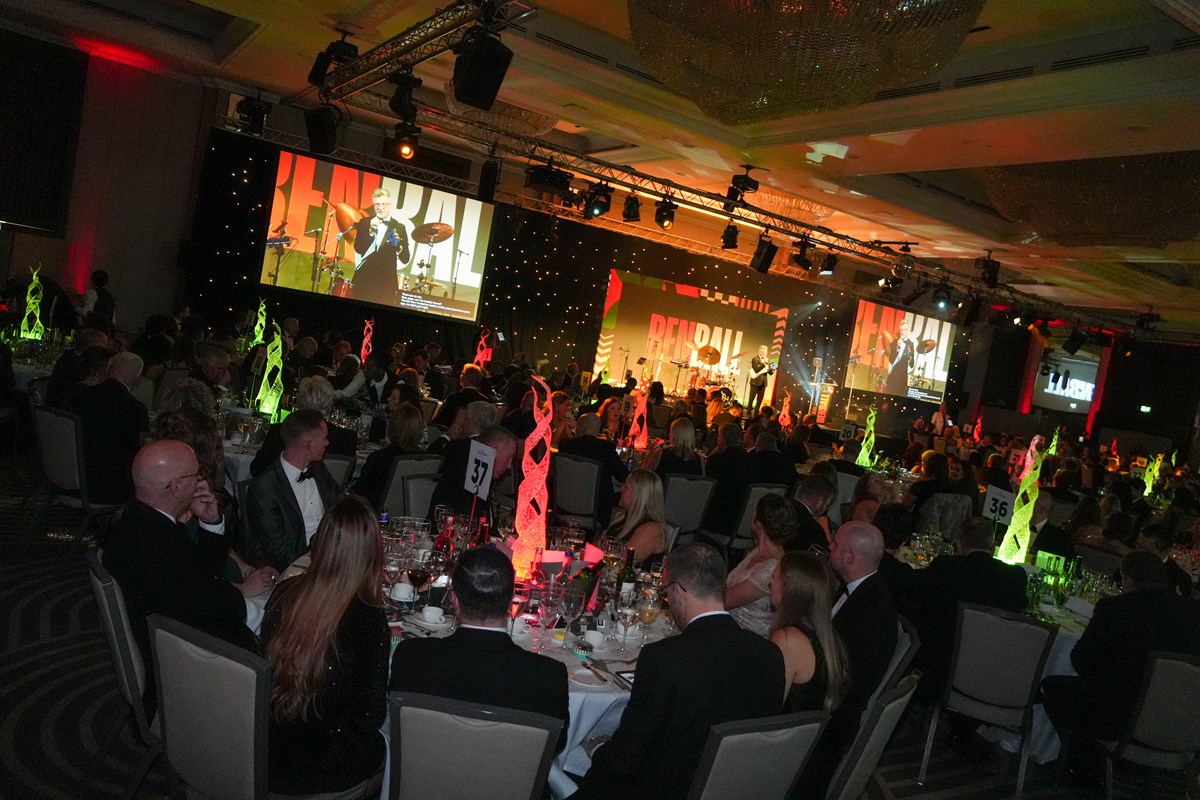 Guests in black tie seated at round tables in a ballroom watching a band on stage, with 'Ben Ball' displayed on large screens.