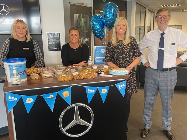 Four colleagues at a Mercedes-Benz dealership standing behind a counter of pastries with a donations bucket, Ben bunting and blue balloons for Ben's Big Breakfast.