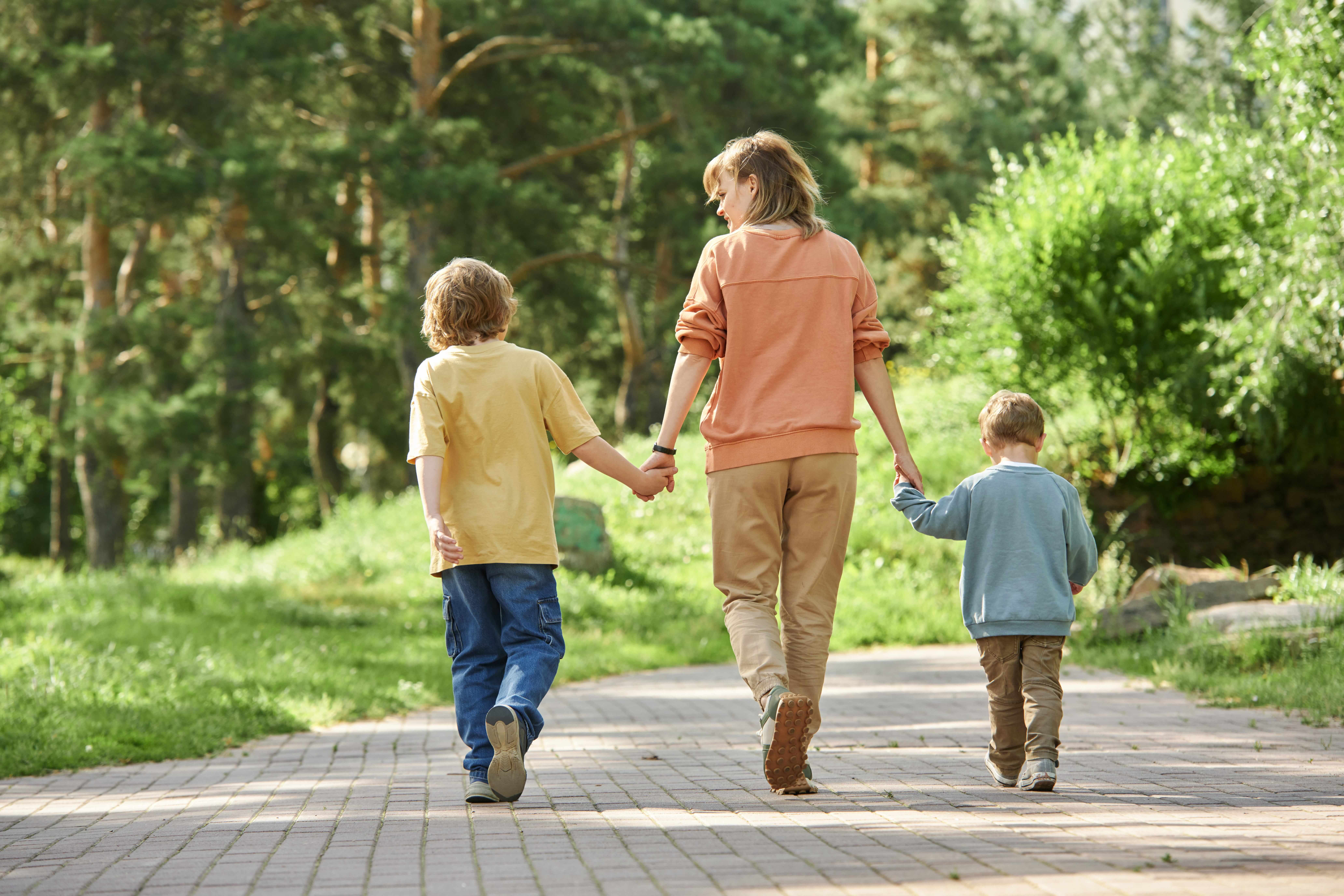 Full length back view of mother with two boys holding hands and walking away from camera in green park