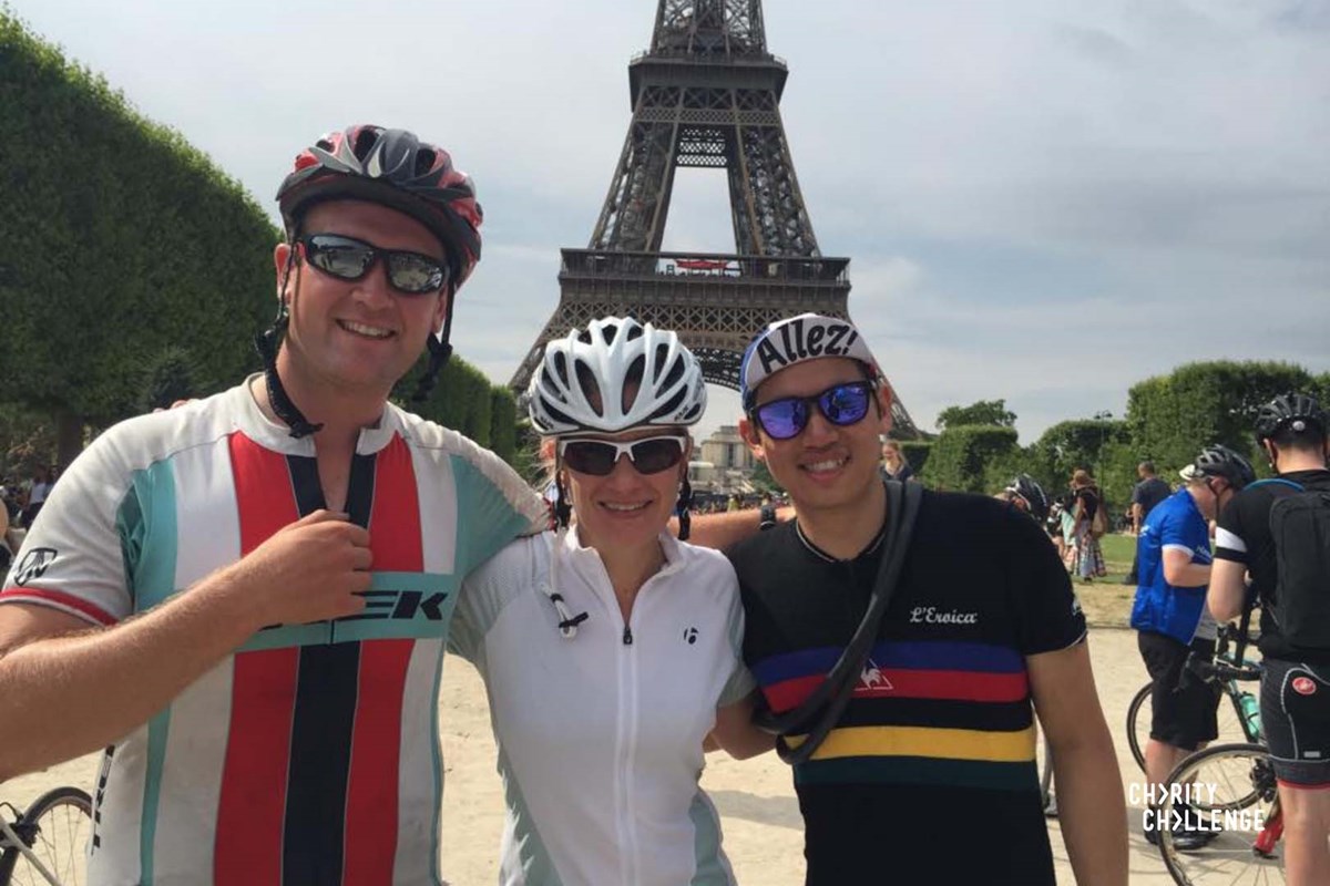 Three cyclists in helmets and cycling kit pose arm in arm in front of the Eiffel Tower at the end of their ride.