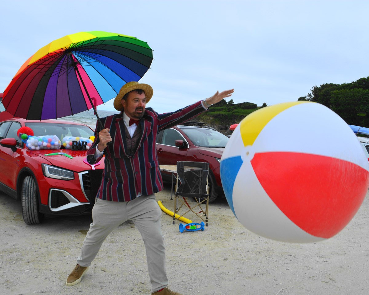 A man in a striped blazer, bow tie and straw boater holds a rainbow umbrella on a beach, batting a giant beach ball, with cars parked behind.