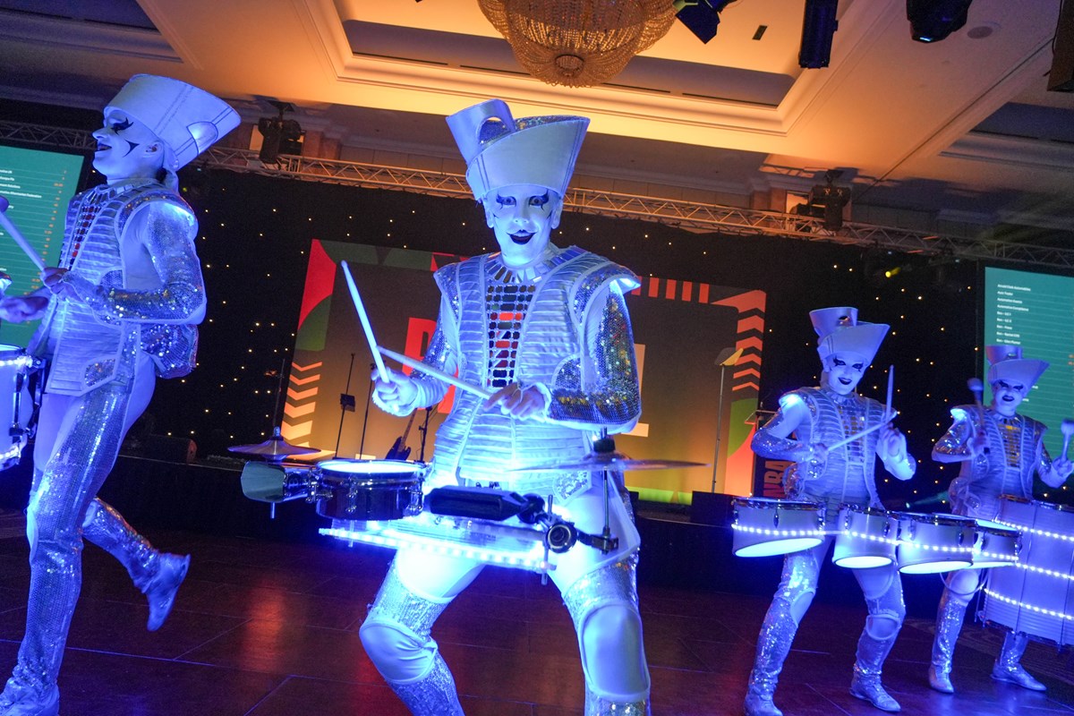 Performers in silver costumes and white face paint play illuminated drums under blue lighting at the Ben Ball.