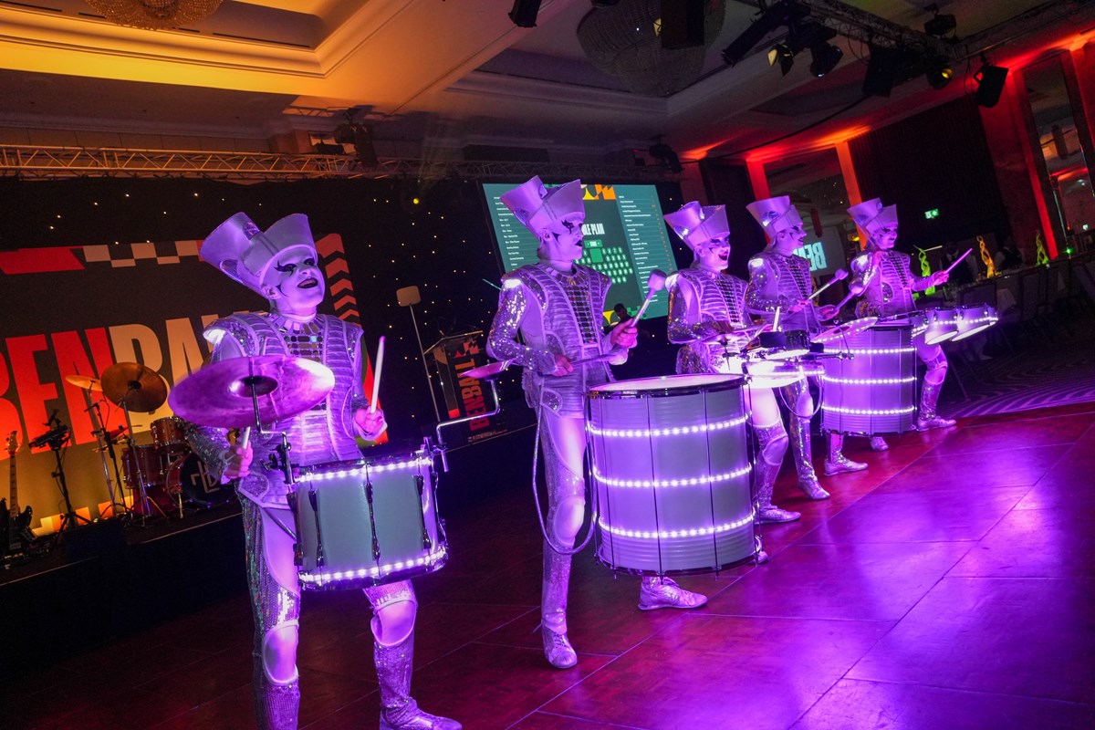 Drummers in silver costumes and tall hats perform with LED-lit drums under purple lighting at the Ben Ball.