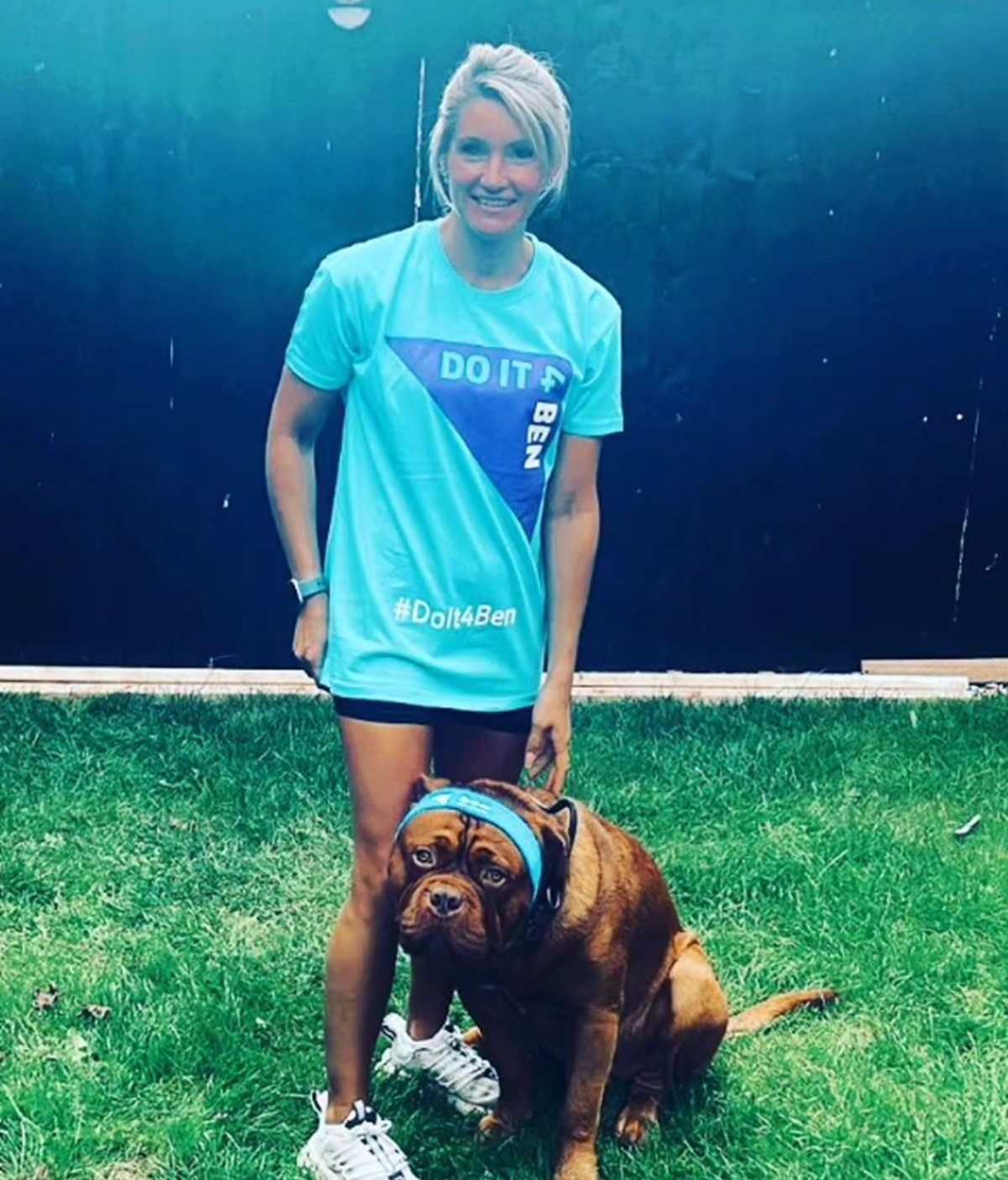 Woman in a turquoise 'Do It 4 Ben' #DoIt4Ben T-shirt standing on grass beside a large brown dog wearing a blue bandana.