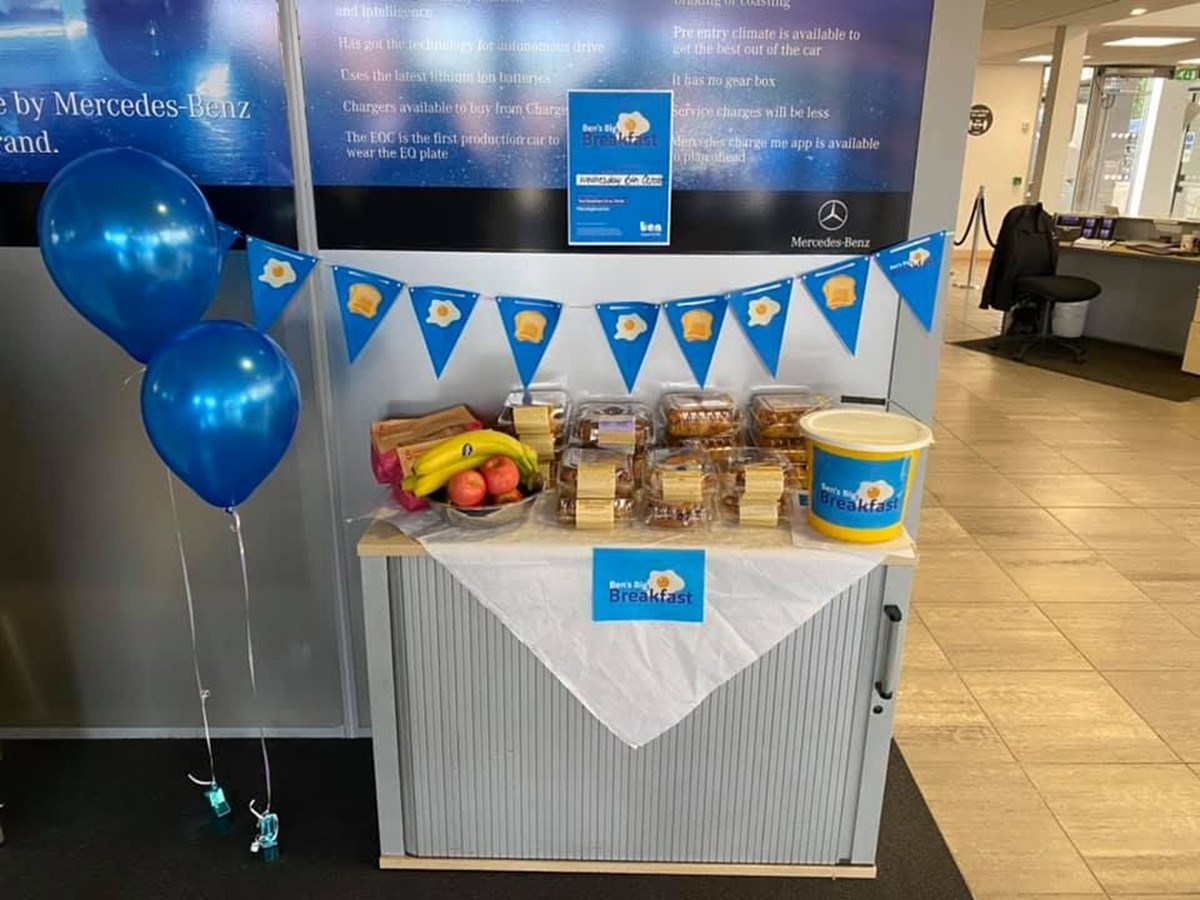 A Ben's Big Breakfast stall in a Mercedes-Benz showroom, with bunting, blue balloons, a collection bucket and a table of cakes and fruit.