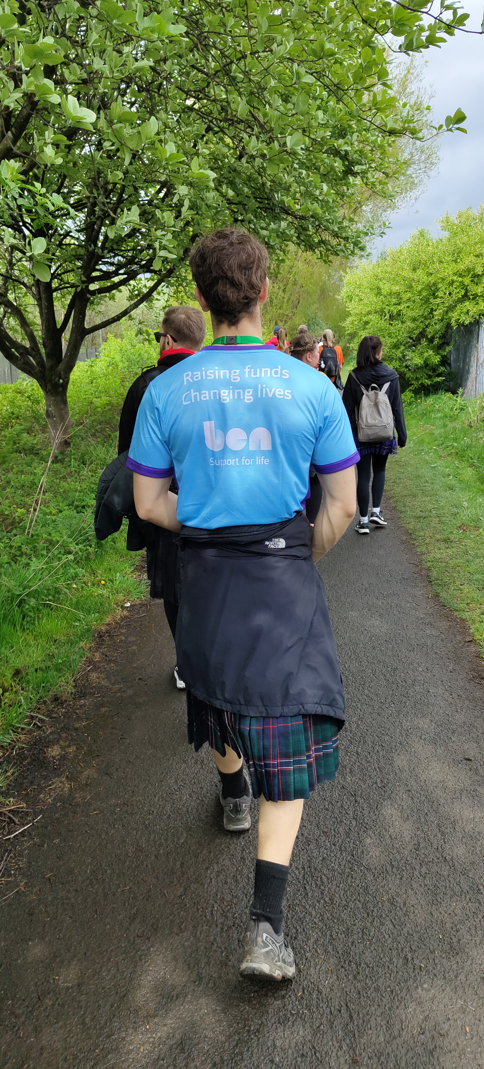 Man walking on the kiltwalk in Glasgow - with a blue t-shirt with white writing which reads Raising funds changing lives and kilt on.