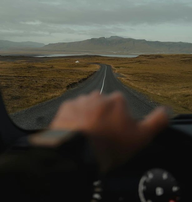 View through a windscreen of a driver's hand on the steering wheel, facing an empty road across open moorland towards distant hills.