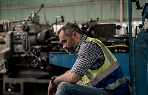 A man in a hi-vis vest sits alone in a workshop, head down and looking downcast.