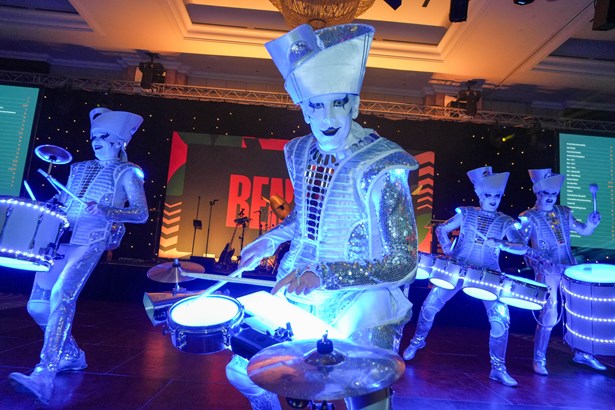 Four performers in silver sequinned costumes and tall hats play illuminated drums on stage at the Ben Ball.