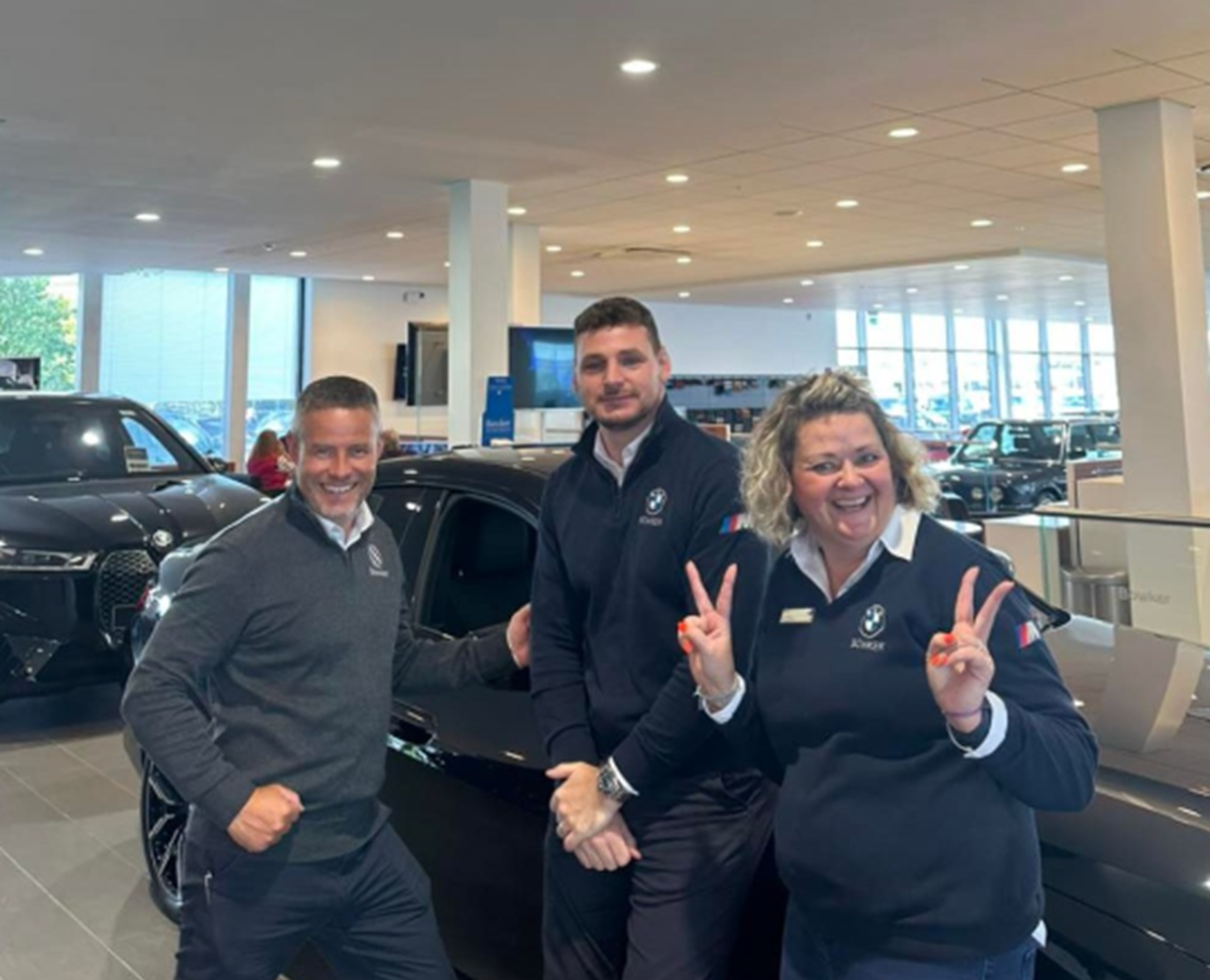 Three car dealership colleagues in branded uniforms smiling and posing with peace signs in a showroom beside a black car.