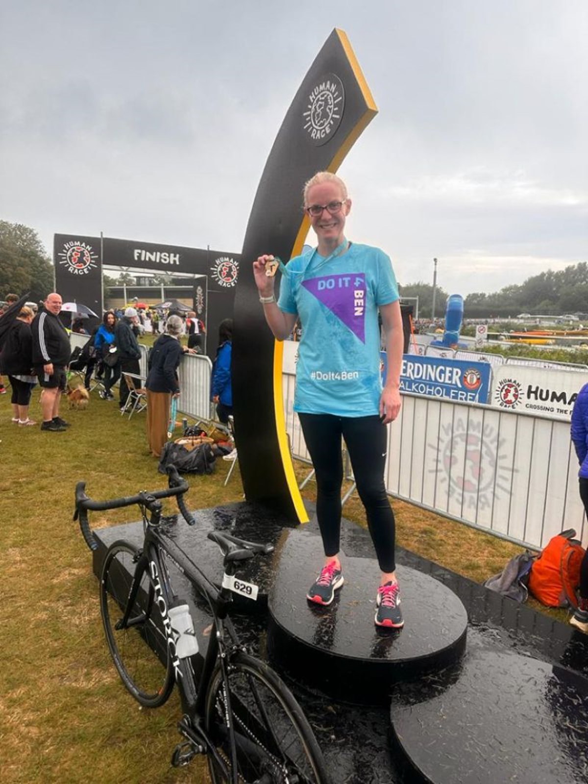 A woman in a 'Do It 4 Ben' T-shirt stands on the winner's podium holding up her medal beside her bike, in front of a triathlon finish line.