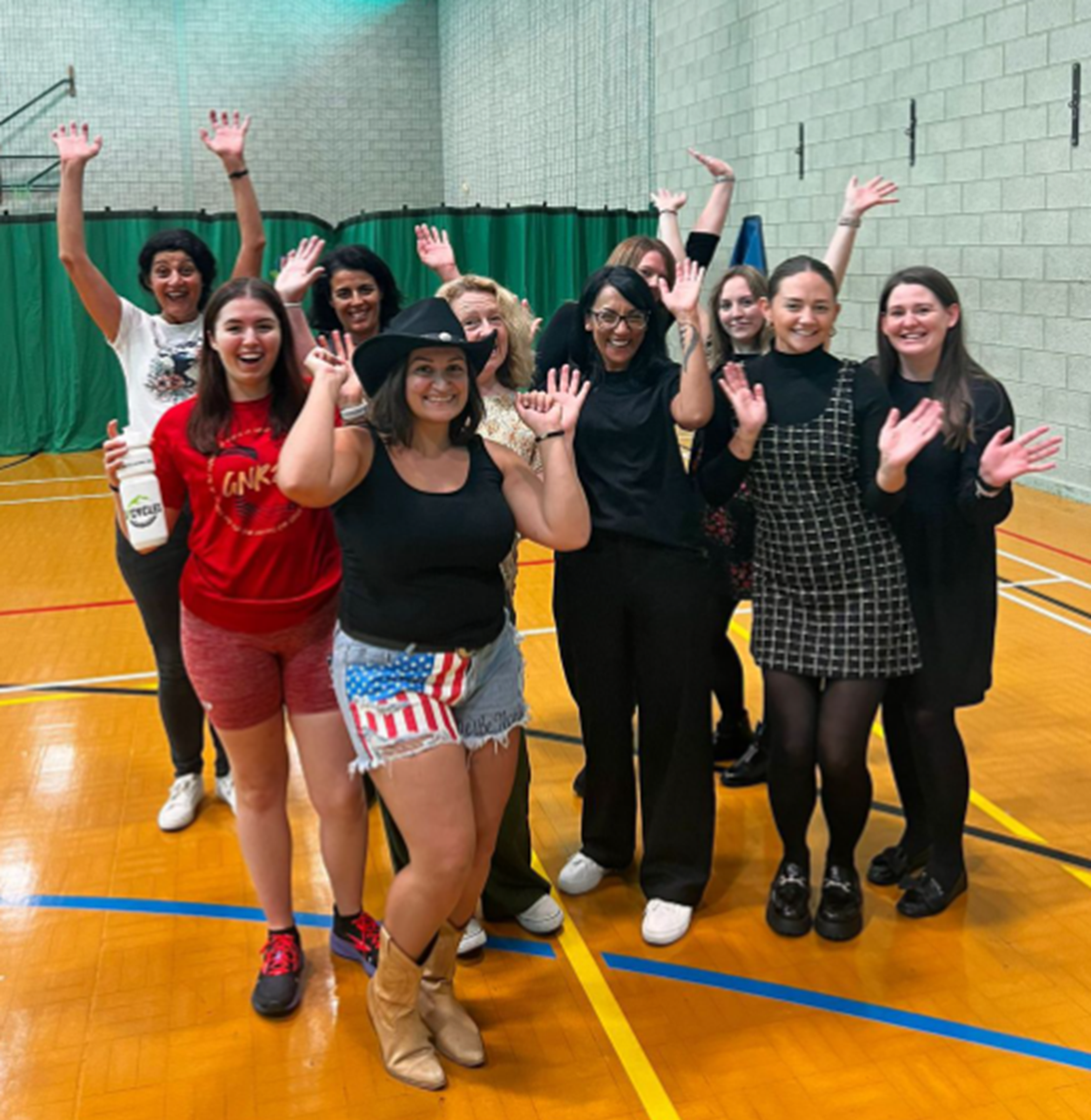 A group of women in a sports hall waving and smiling, some in cowboy boots and hats.