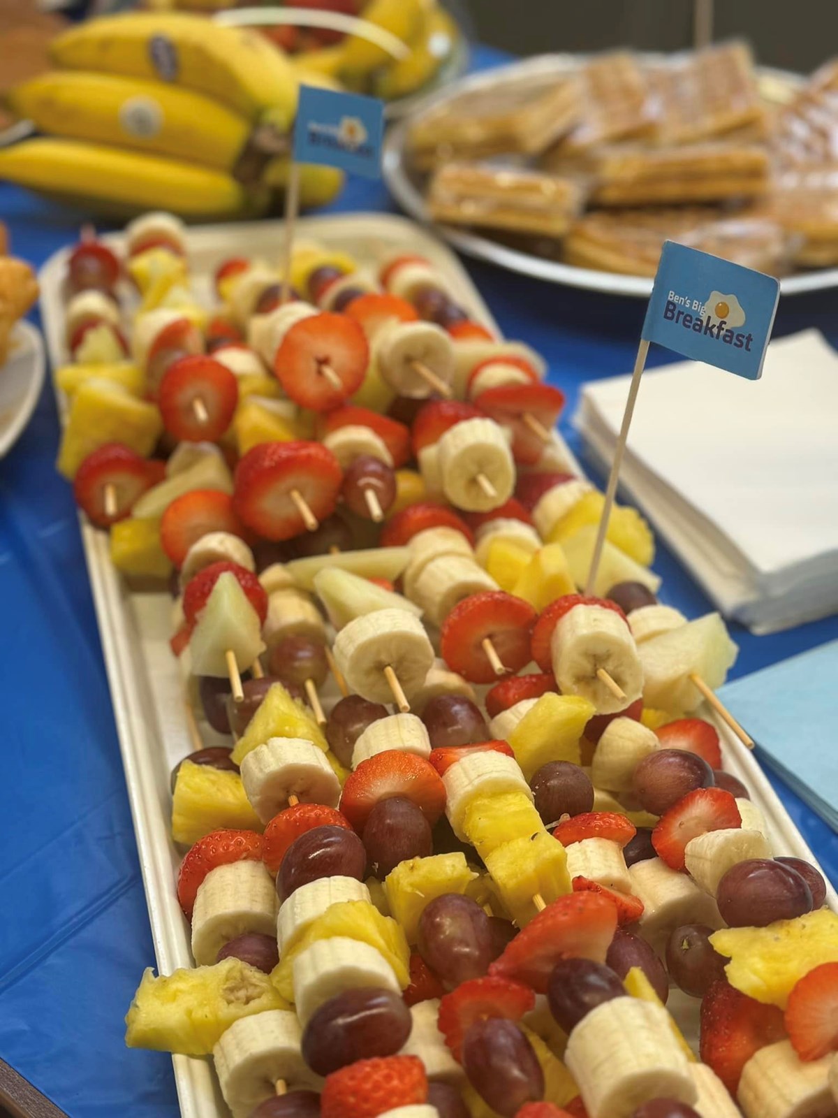 Platter of fresh fruit skewers with strawberries, banana, grapes and pineapple, marked with 'Ben's Big Breakfast' flags, beside bananas and pastries.