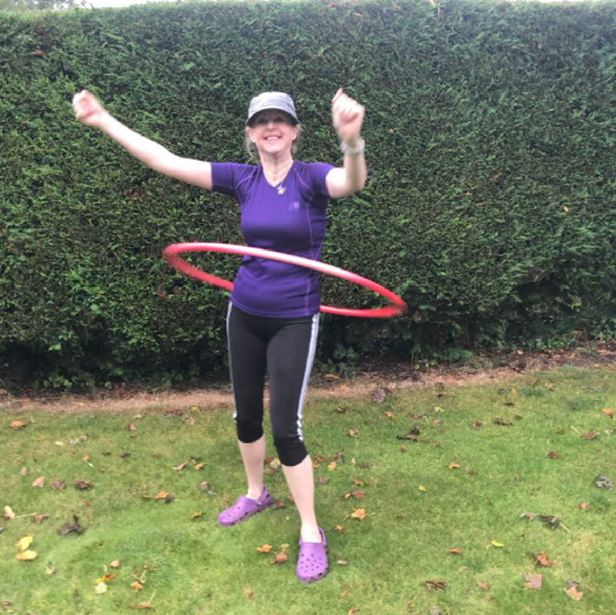 Woman in a purple T-shirt and cap spinning a red hula hoop on a lawn in front of a hedge, arms raised in celebration.