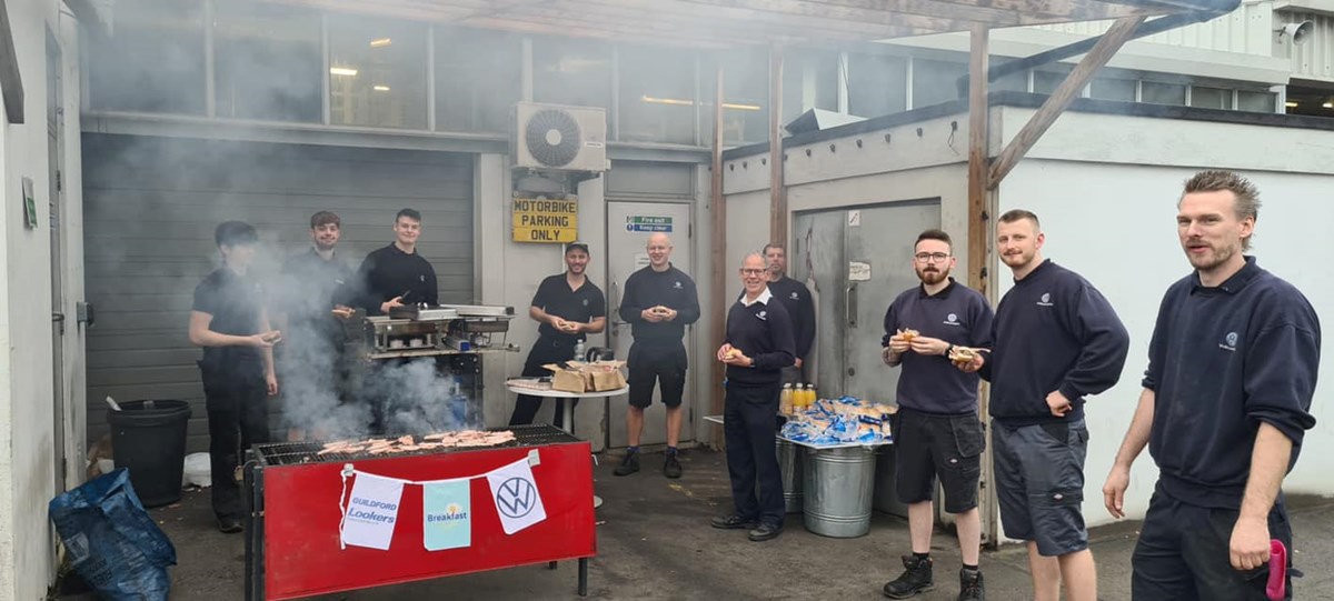 Dealership colleagues gathered outside around a smoking barbecue with bacon rolls, with signs reading 'Guildford Lookers', 'Big Breakfast' and the VW logo.