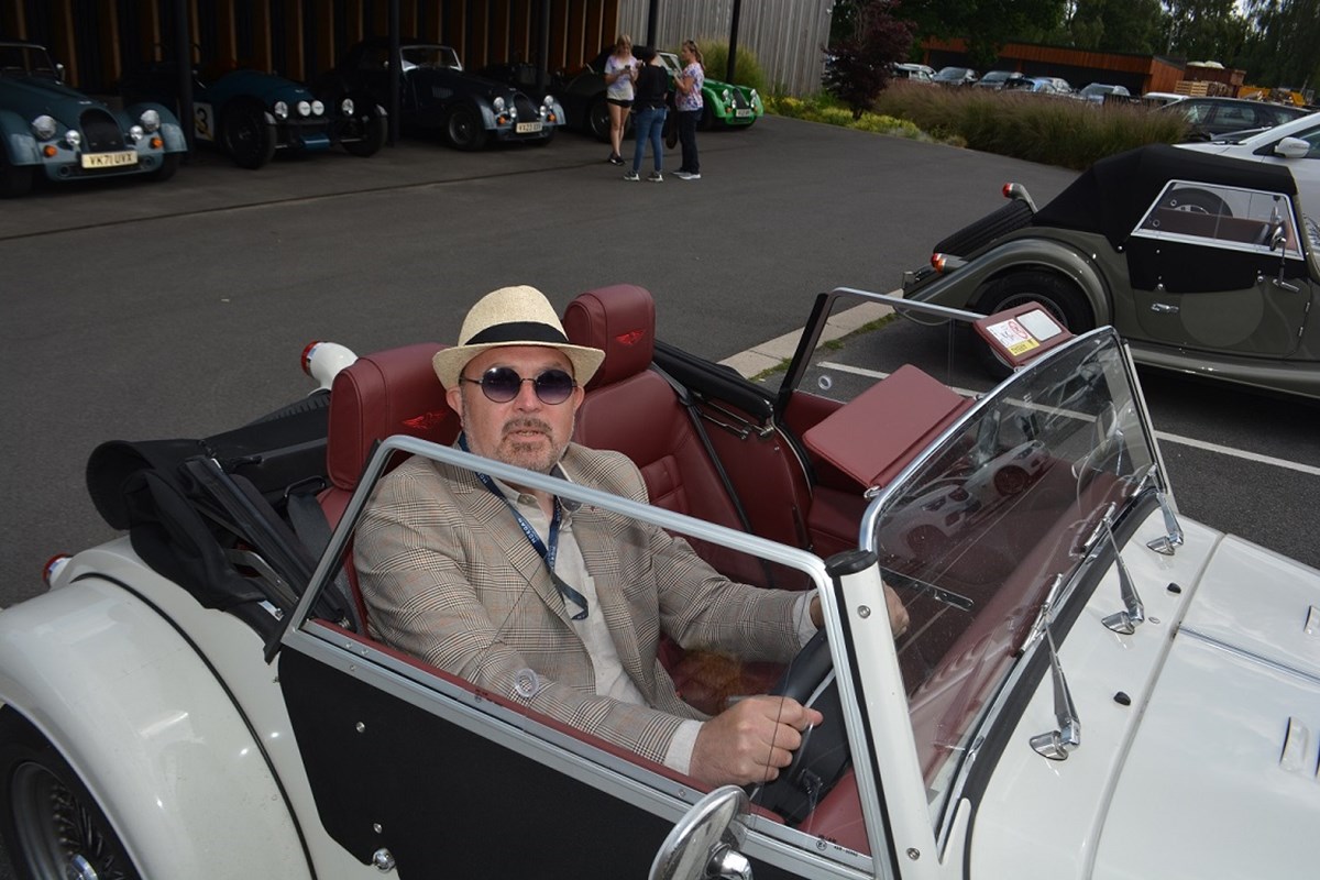 A man in a straw hat, sunglasses and checked jacket sits at the wheel of a white open-top classic sports car, with more classic cars parked behind.