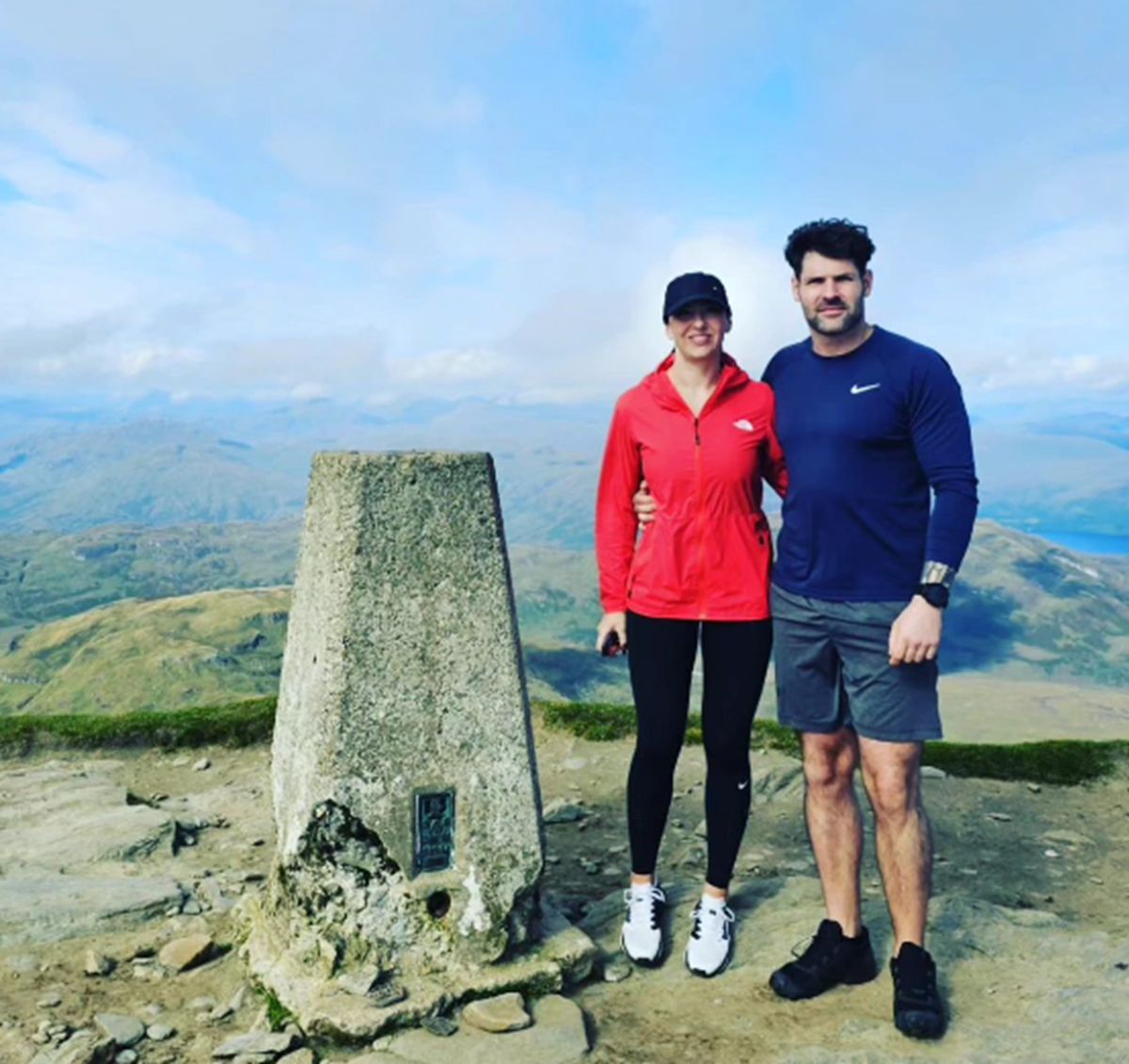 A woman in a red jacket and a man in a navy top stand arm in arm beside a stone trig point at a mountain summit, with hills and lochs behind them.