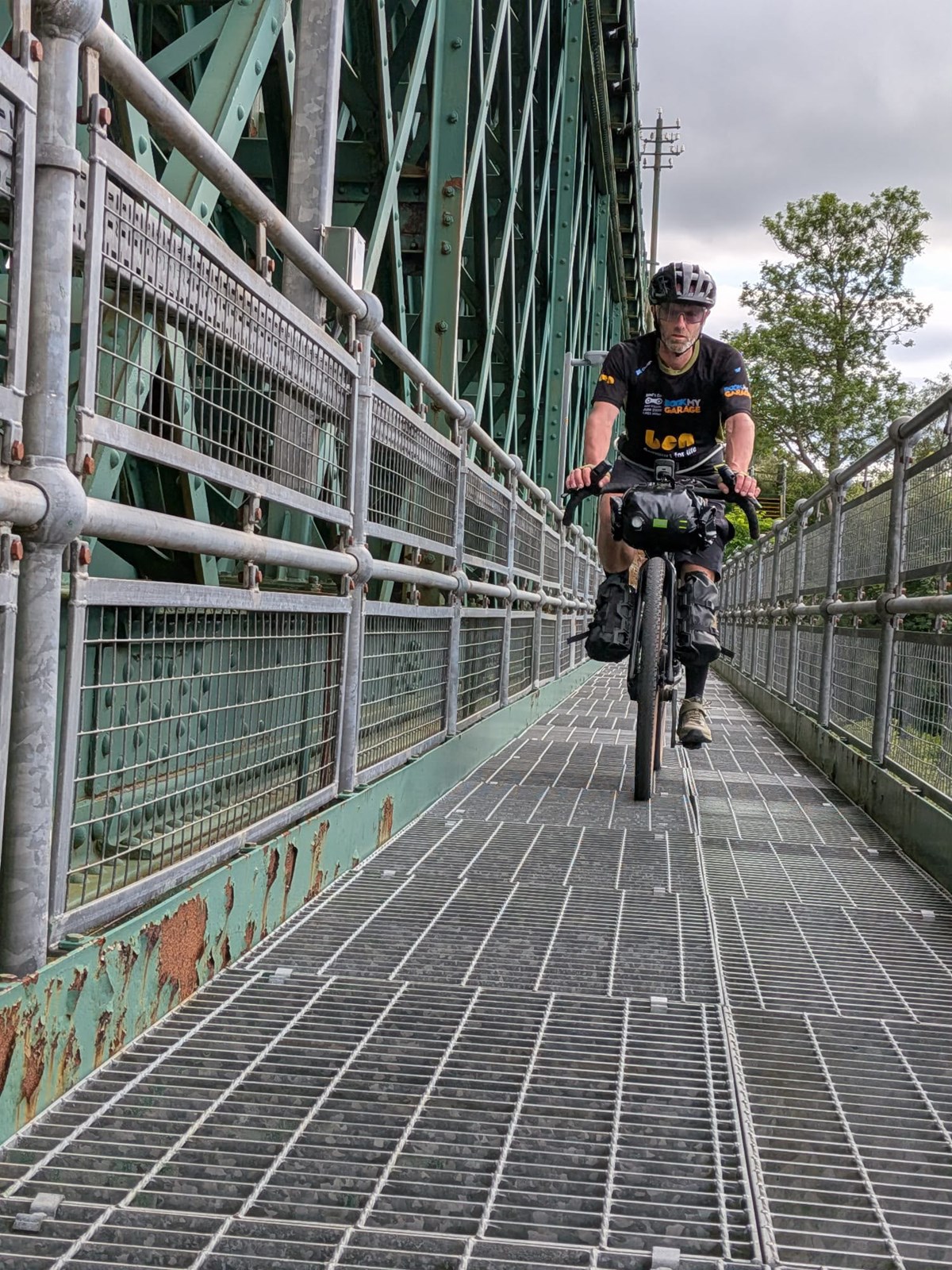 Cyclist in a branded jersey with loaded bikepacking bags riding across a metal walkway on a green iron bridge.