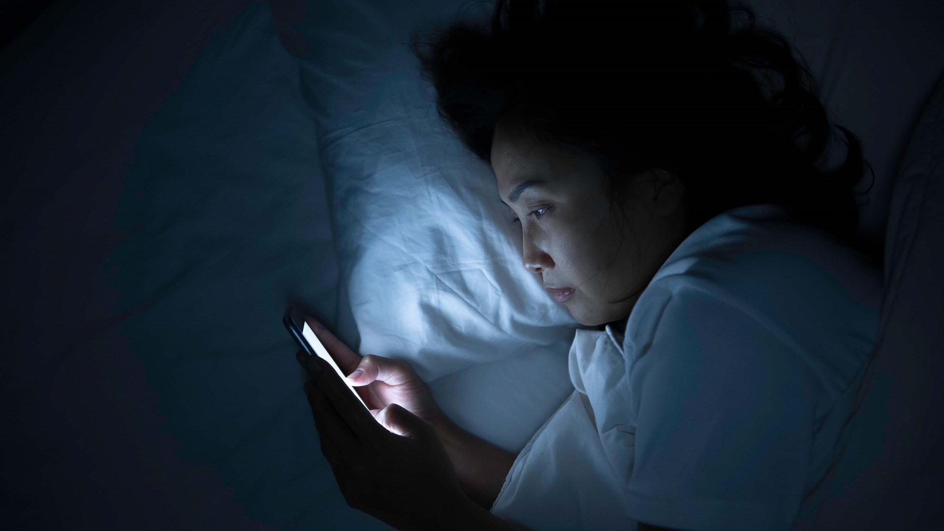 Woman lying awake in bed at night, her face lit by the screen of her phone.