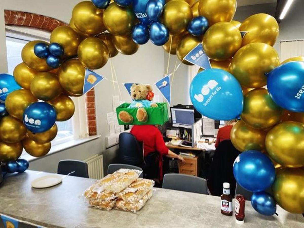An office decorated with a gold and blue balloon arch and Ben 'Support for life' balloons and bunting, with a teddy bear and trays of breakfast rolls and sauces laid out on a table.