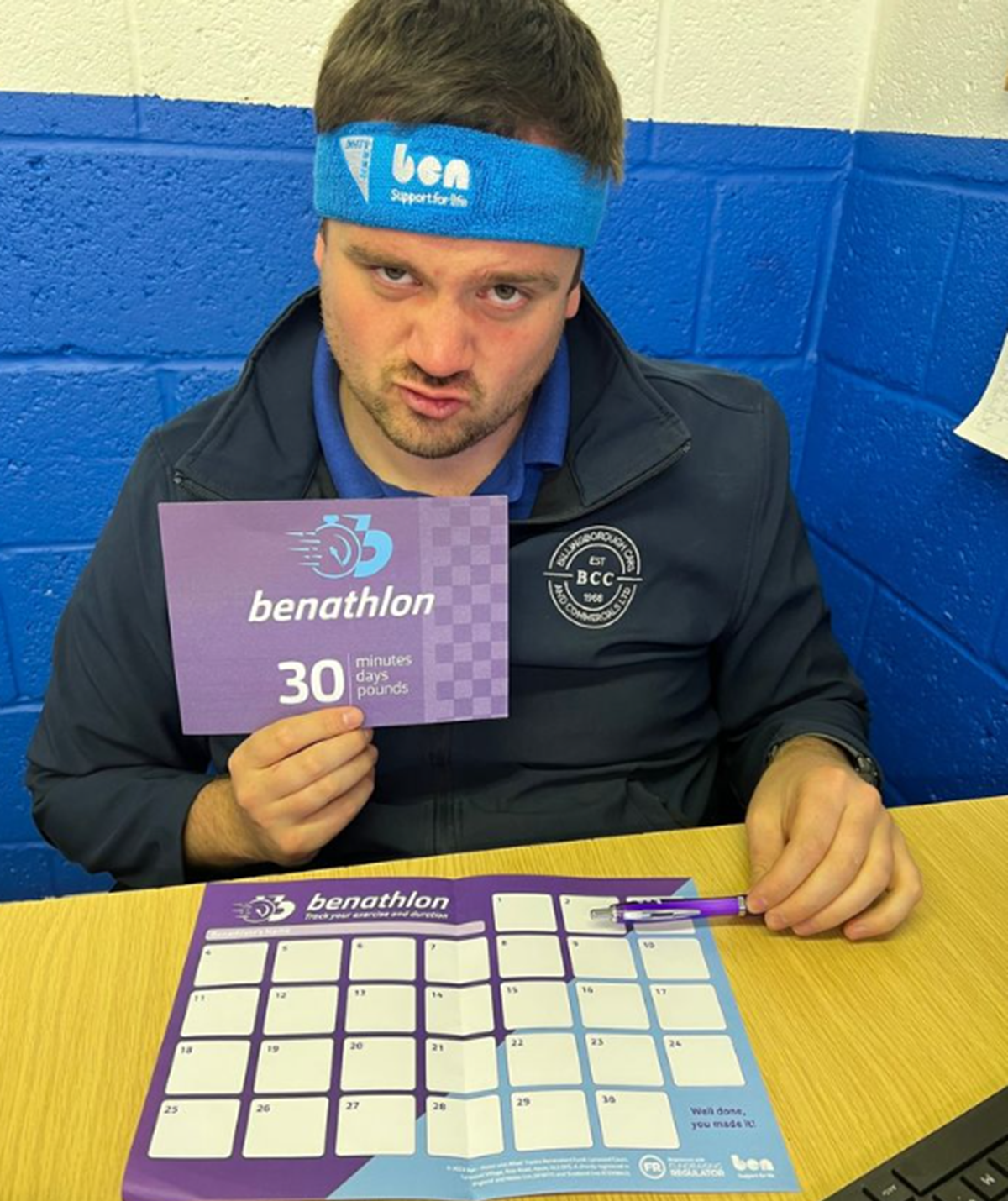 A man wearing a blue Ben headband holds up a 'Benathlon 30 minutes, days, pounds' card, with a Benathlon exercise tracker chart on the desk in front of him.