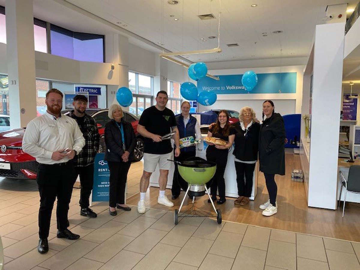 Dealership team gathered around a barbecue in a Volkswagen showroom with blue balloons, holding buns and sausages for Ben's Big Breakfast.