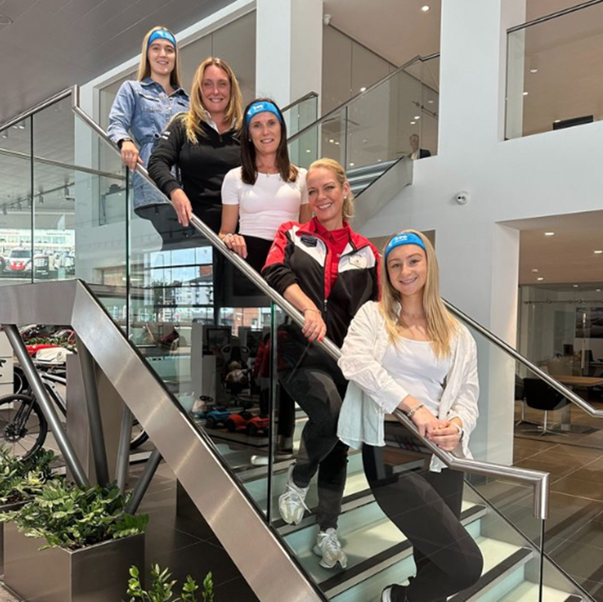 Five colleagues wearing blue headbands pose on the glass staircase of a car showroom.