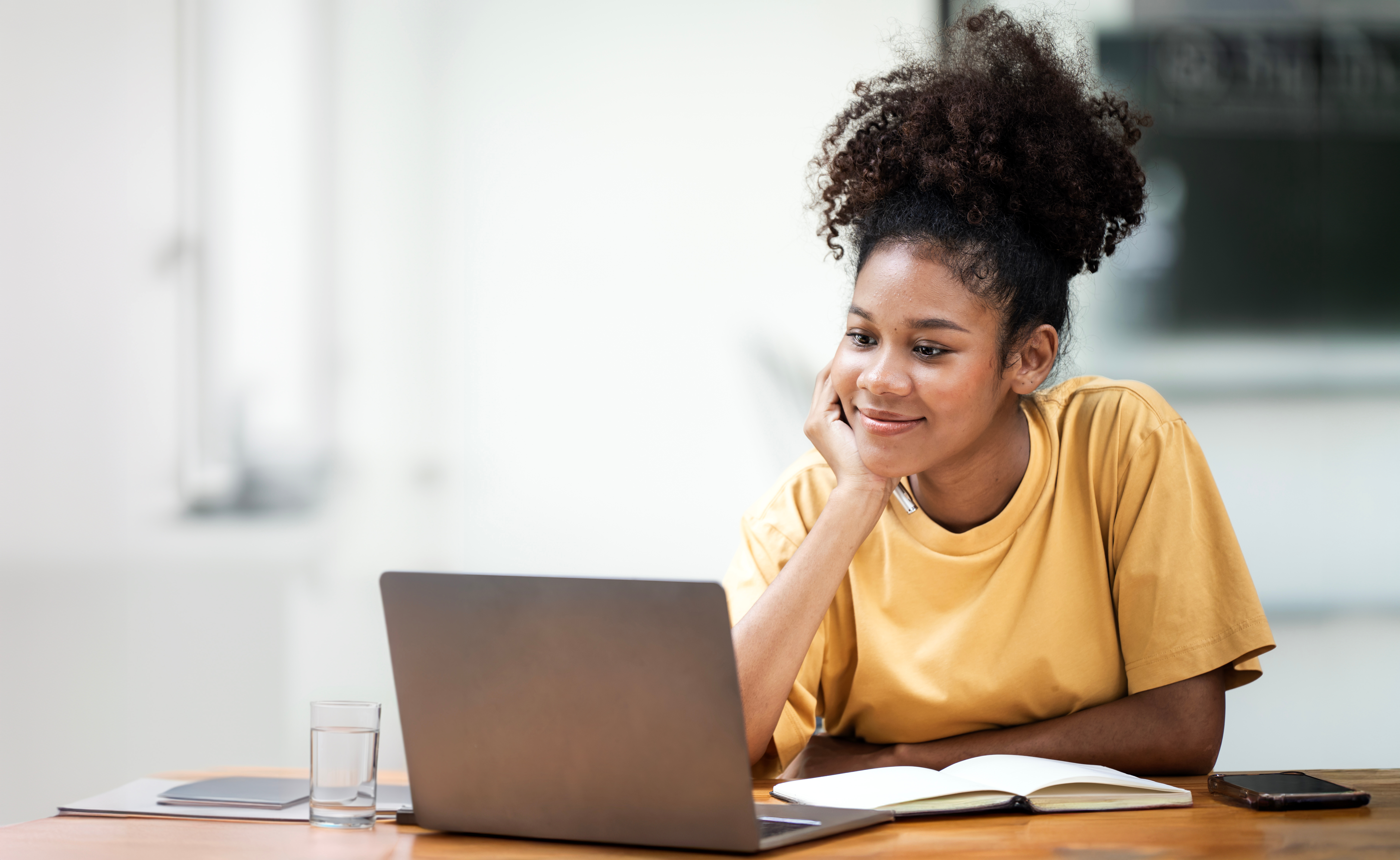 Smiling young black woman student sitting at desk studying using using laptop at a desk. Wearing a bright yellow t-shirt.