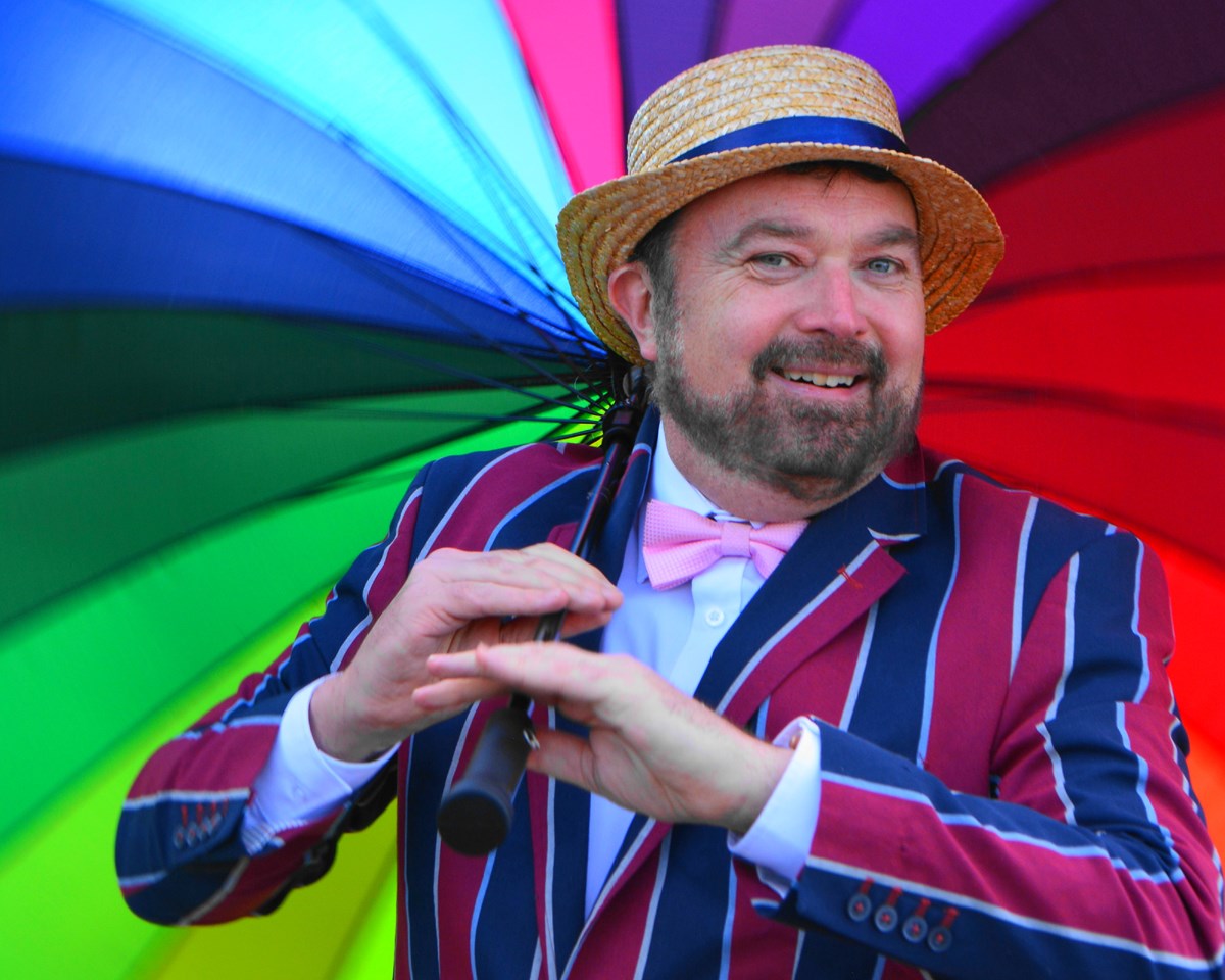 Smiling man in a straw boater hat, striped blazer and pink bow tie holding a rainbow-coloured umbrella.
