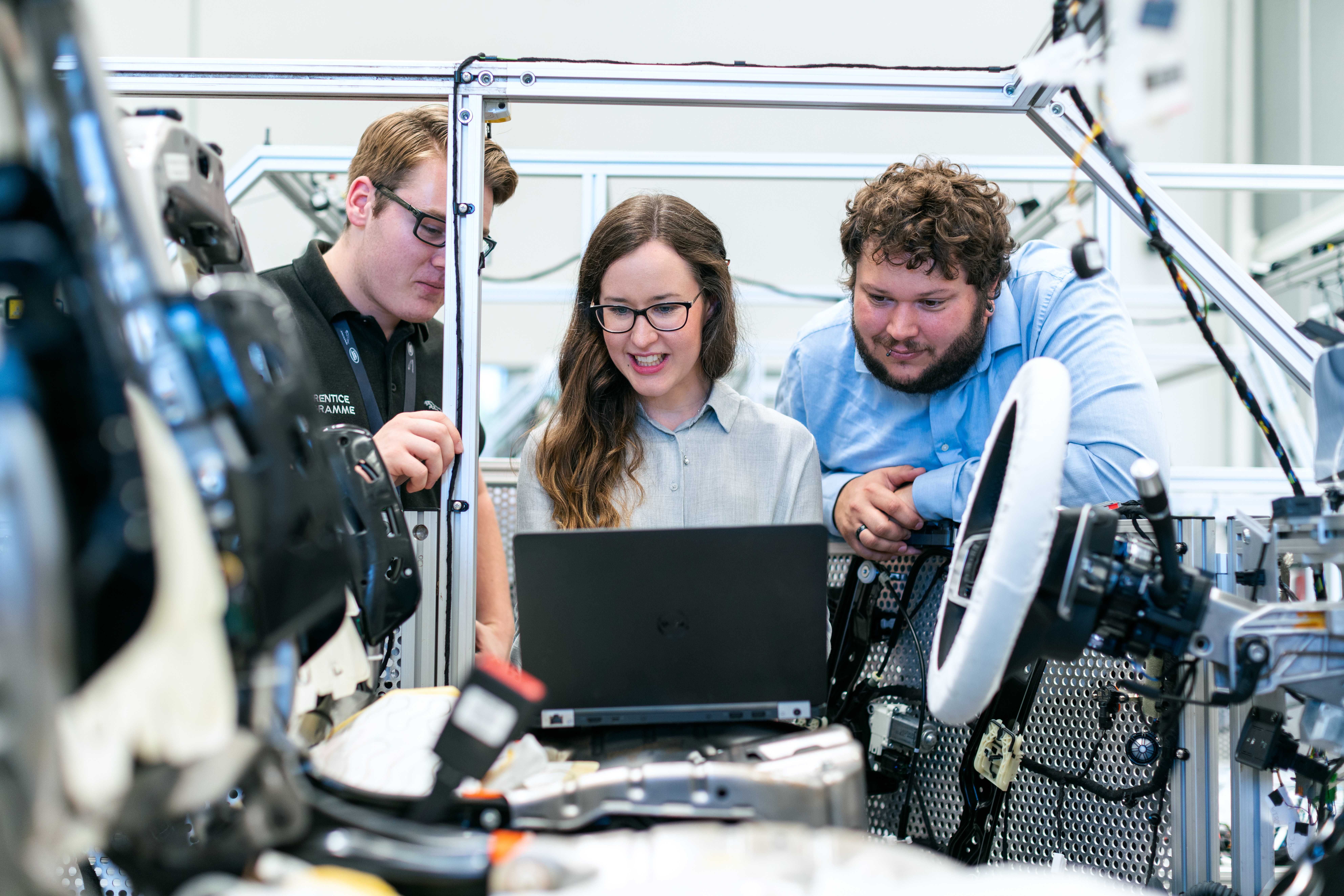 Three automotive engineers looking at a laptop together beside vehicle components in a workshop.