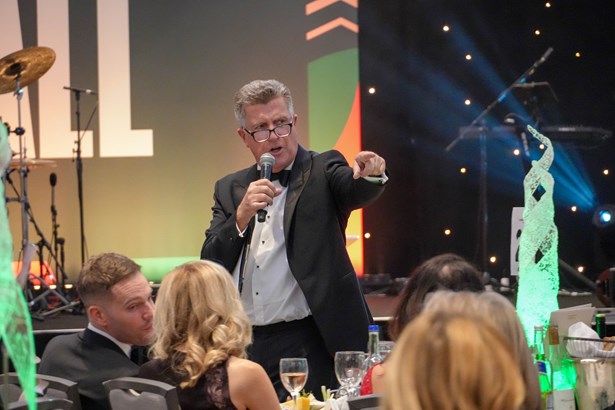 An auctioneer in black tie holds a microphone and points into the audience during a charity ball auction.