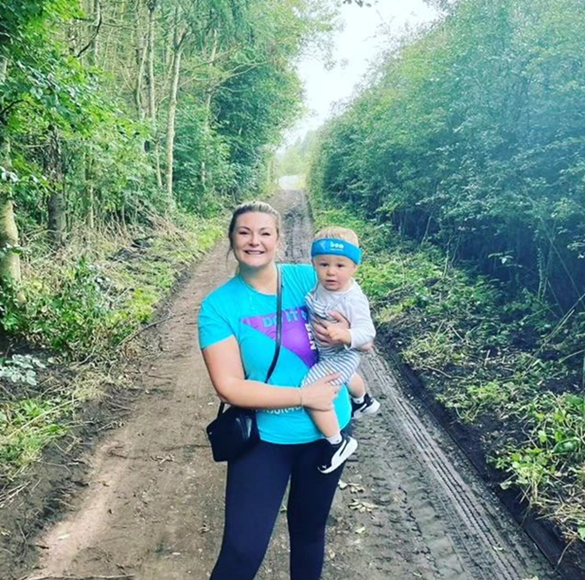 A woman in a Ben charity T-shirt carrying a toddler wearing a blue Ben headband along a muddy woodland track.