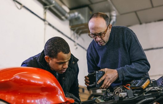 An older mechanic shows a car part to a younger colleague as they work under the bonnet of a red car in a workshop.
