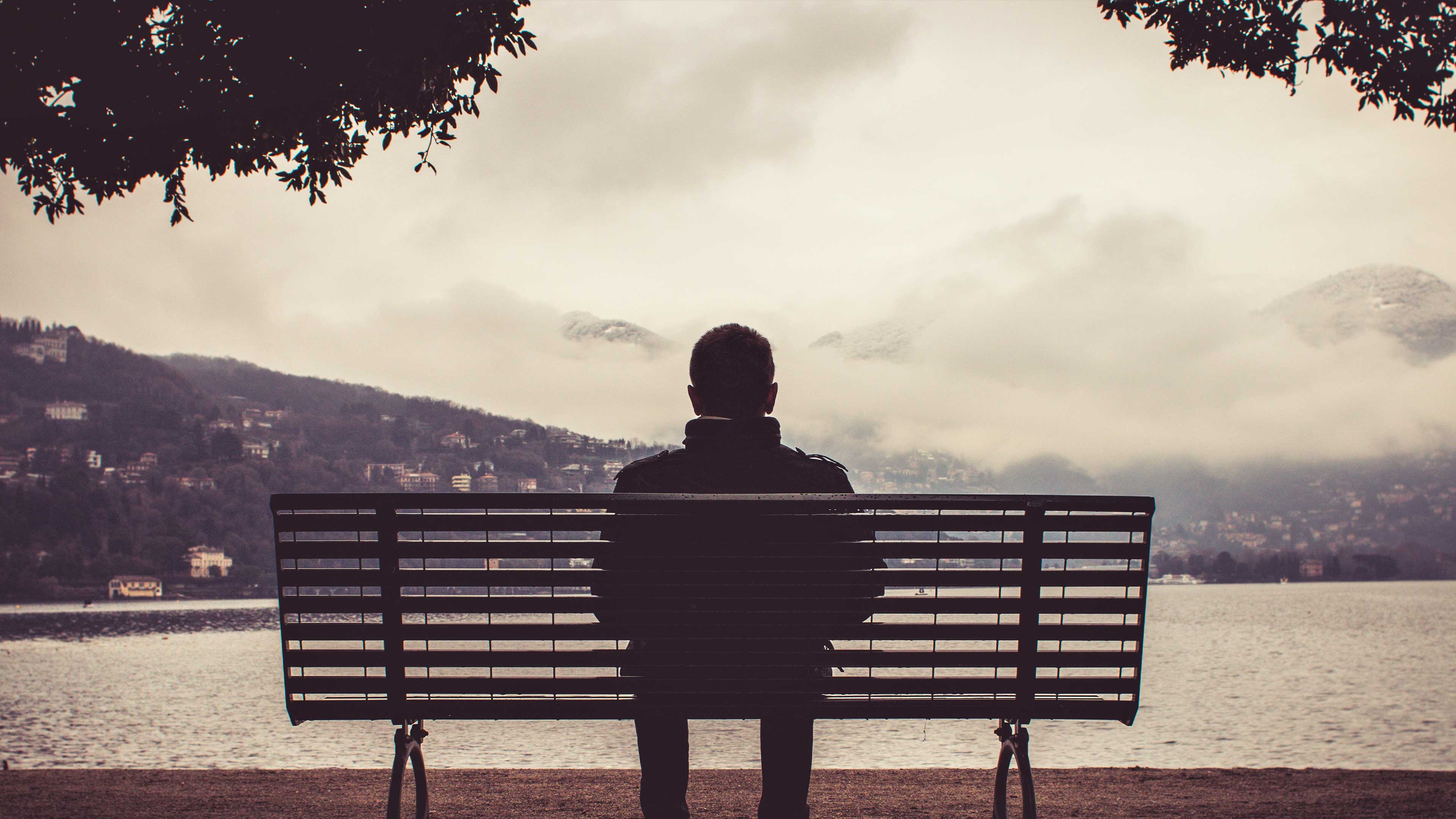 A man sitting alone on a bench, seen from behind, looking out across a lake towards misty hills.