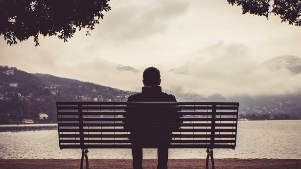 A man sitting alone on a bench, seen from behind, looking out across a lake towards misty hills.