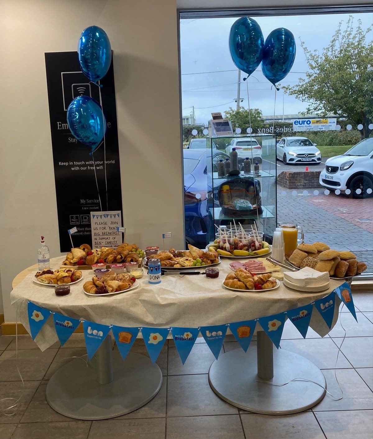 Car showroom table laid with pastries, croissants, bread rolls, fruit and juice, decorated with Ben's Big Breakfast bunting, blue balloons and a donation tin.