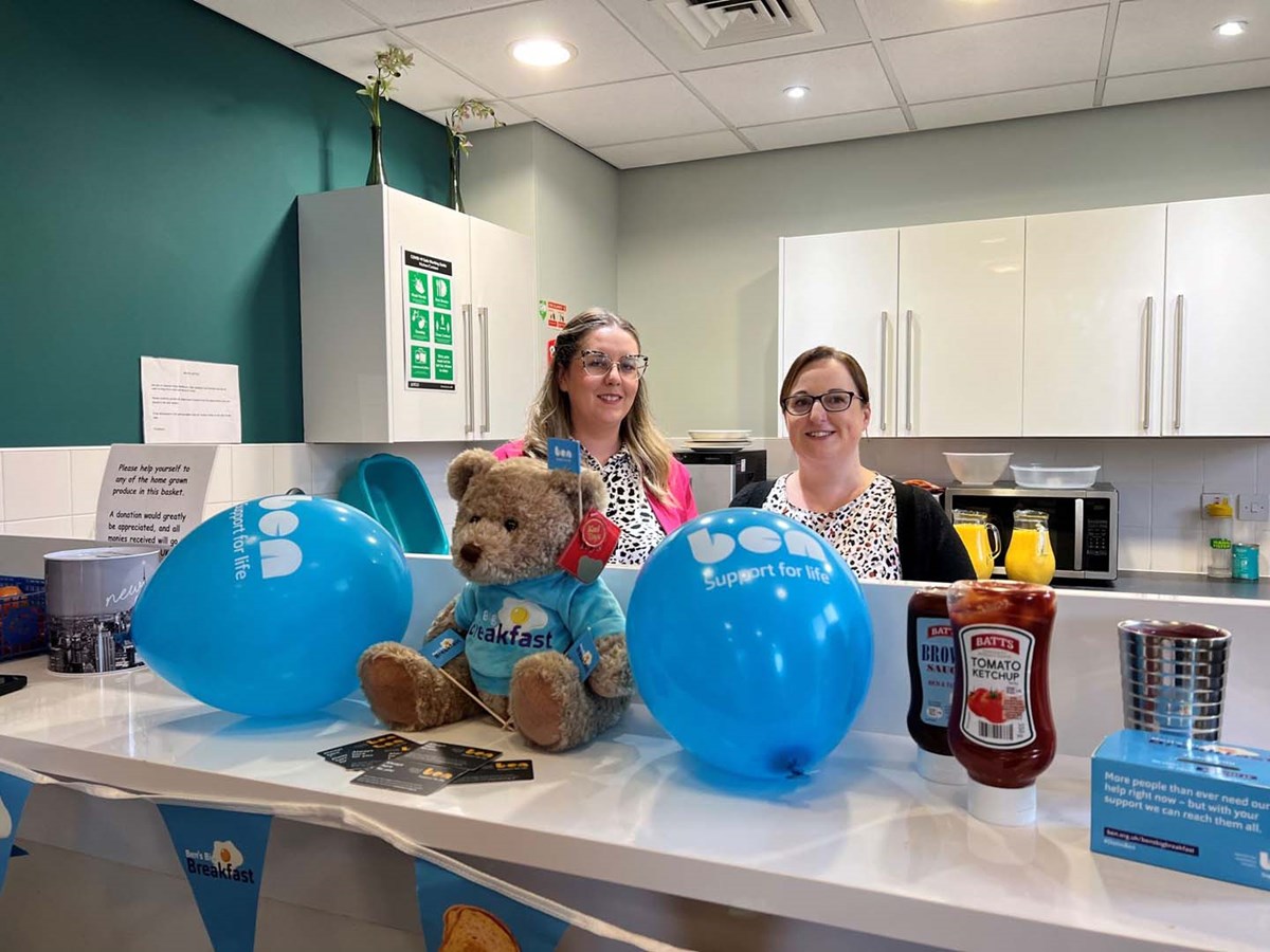 Two women smiling behind a kitchen counter decorated with Ben balloons, bunting and a teddy bear in a Ben's Big Breakfast T-shirt.