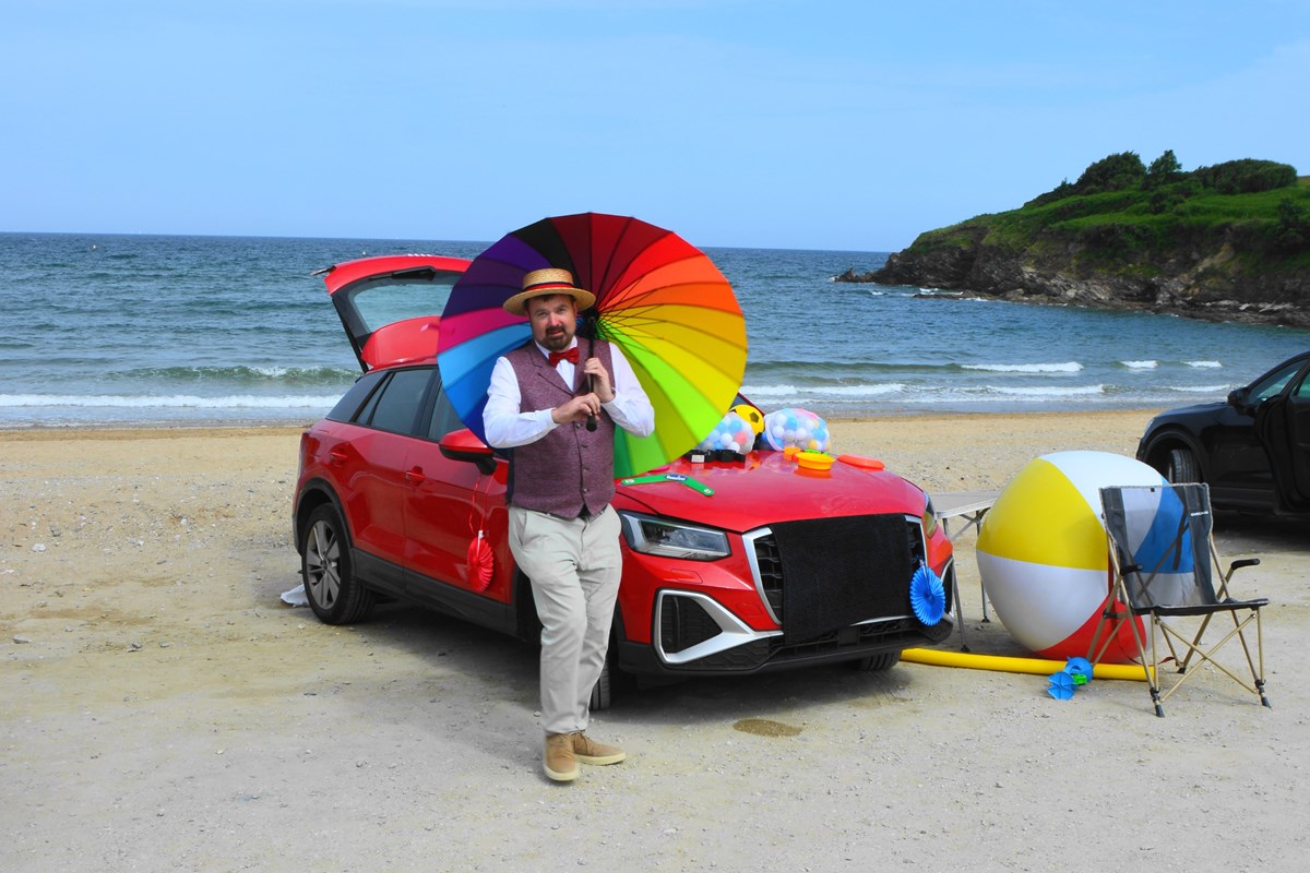 A man in a straw boater, waistcoat and bow tie holding a rainbow umbrella beside a red car on a beach, with a giant beach ball and deckchair.