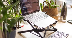 Laptop on an adjustable stand so it becomes eye level. A clean desk with additional keyboard and mouse. Several plants decorate the desk.