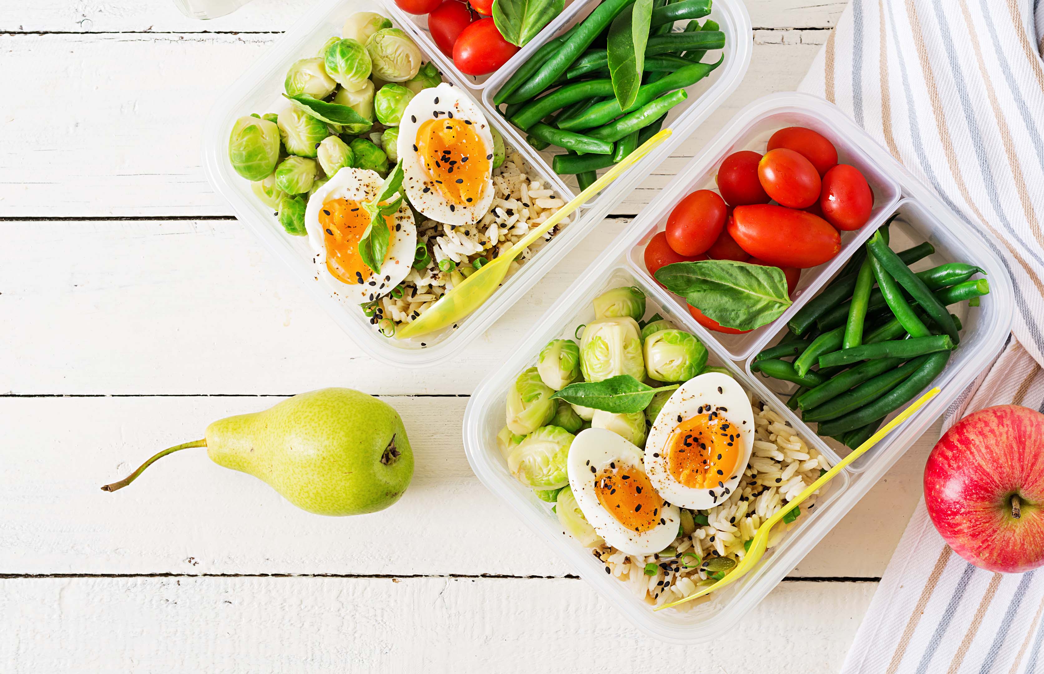 Two lunchboxes filled with colourful healthy foods ready to enjoy on the go. There is a red apple to one side of the boxes, and a green pear to the other.