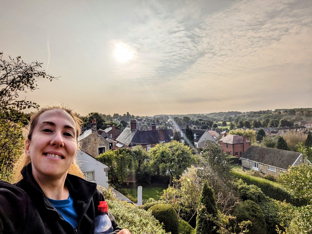 A smiling woman takes a selfie with a water bottle, with a sunlit view over village rooftops and trees behind her.