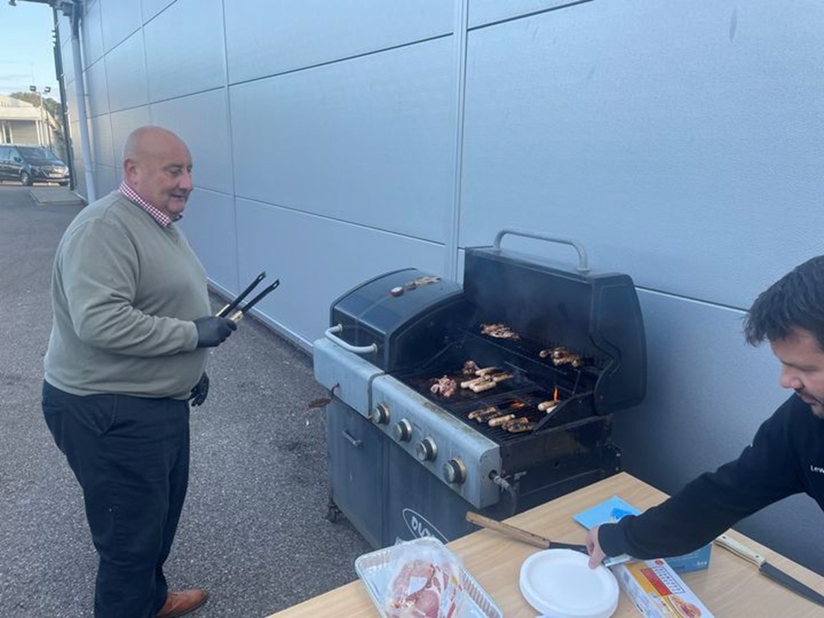 A man holding tongs smiles beside a barbecue grilling sausages and bacon outside a dealership, while a colleague sets out plates on a table.