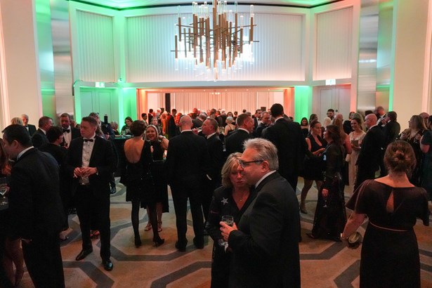 Guests in black tie mingling with drinks in a hotel foyer lit with green uplighting beneath a large chandelier at the Ben Ball drinks reception.