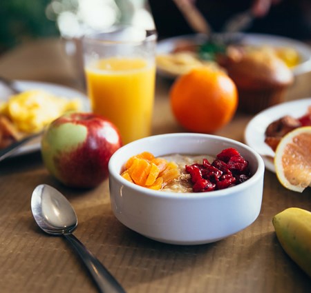 A white bowl full of fruit with a spoon next to it sitting on a table. The rest of the table contains more fruit and other healthy breakfast foods  